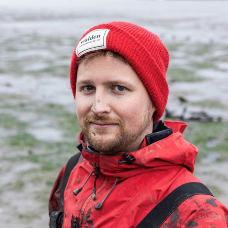 Ollie is standing beside a seagrass meadow. He is wearing a red rain jacket, waders, and a red hat. He has a beard and moustache. Ollie's coat and face have mud on them from fieldwork.